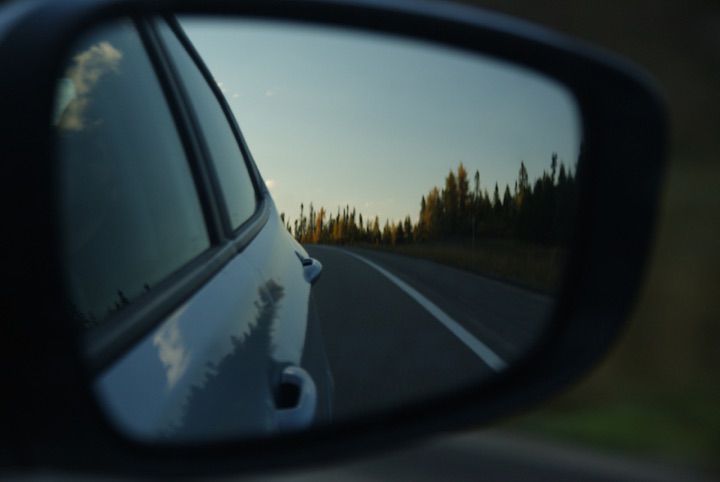View in a car’s rear-view mirror of a road and trees behind the vehicle at dusk.