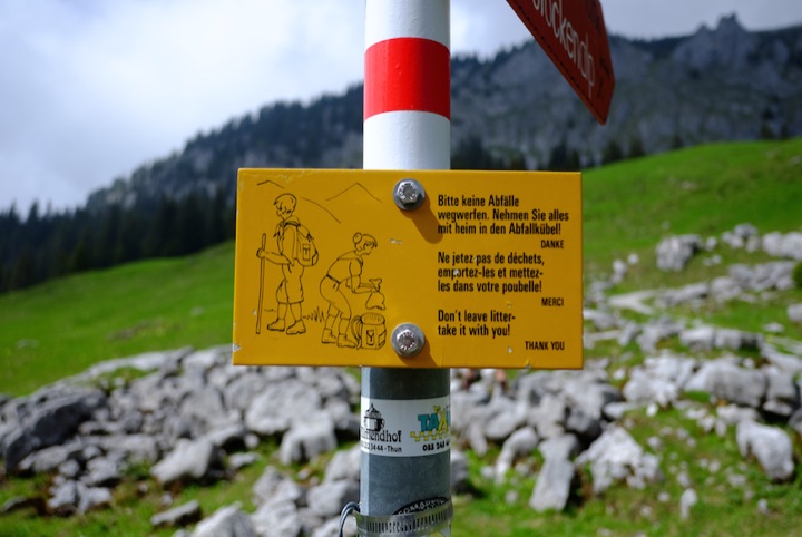 Yellow multilingual litter notice attached to a red-and-white pole in a grassy mountain landscape.