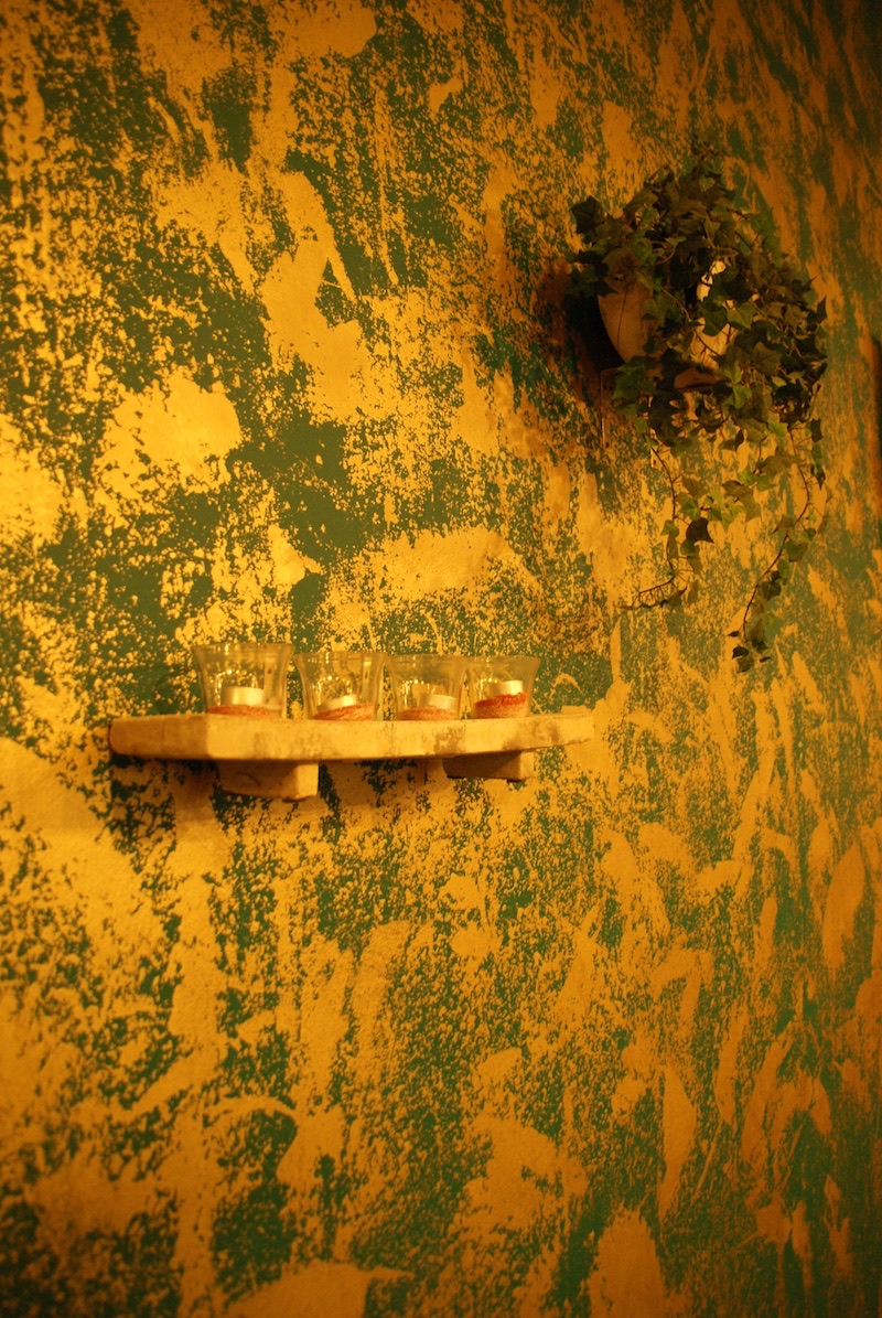 Glass candle holders on a small shelf against a textured yellow-and-green plaster wall, with a potted plant above.