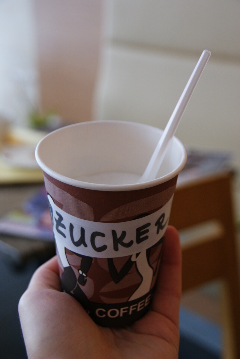 Hand holding a takeaway coffee cup labeled ‘ZUCKER’ with a white plastic spoon.