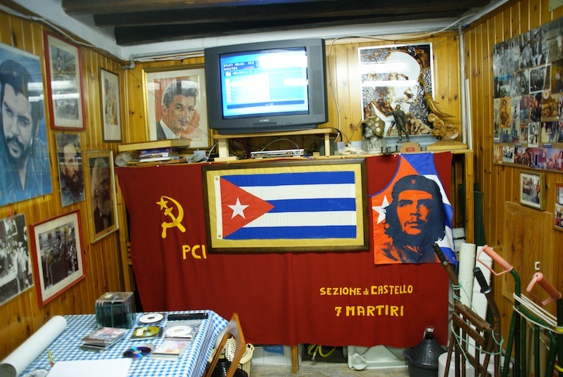 Wood-paneled room displaying framed portraits, a Cuban flag, and a red hammer-and-sickle banner beneath a television.