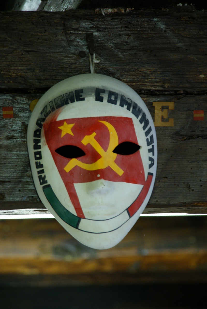 Painted white face mask with a red hammer-and-sickle emblem and Italian lettering against a dark background.