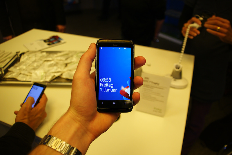 Hand holding a black HTC smartphone with a blue screen; another phone and people appear behind it.