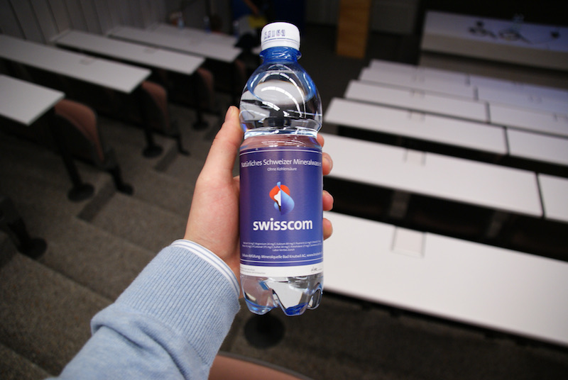 Hand holding a Swisscom mineral-water bottle in a classroom.