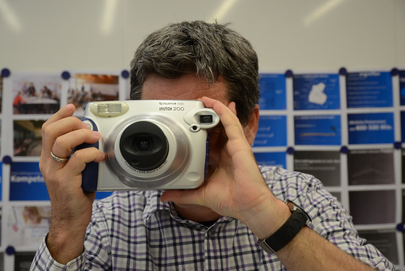 Person holding a large instant camera in front of a board of pinned photographs and blue cards.