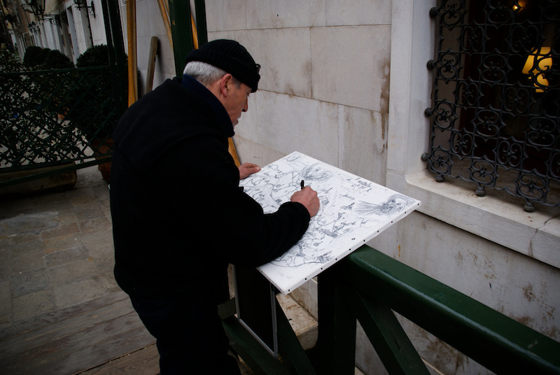 Person in a black coat and cap drawing at an outdoor stand.