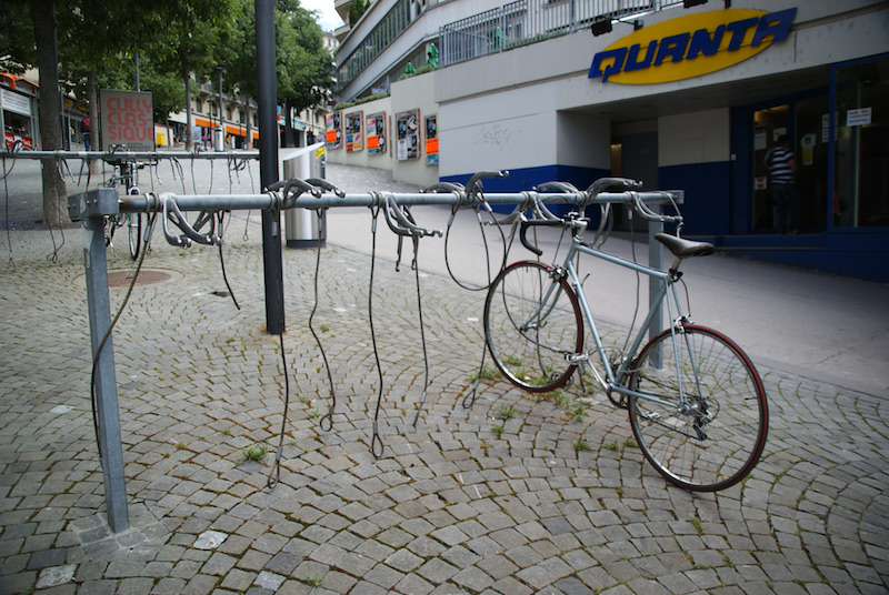 One bicycle locked to a metal rail amid empty locks at a bicycle stand outside a store.