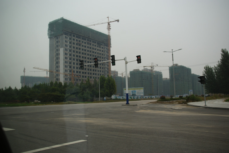 High-rise buildings under construction with cranes beside a wide road and traffic lights in hazy weather.