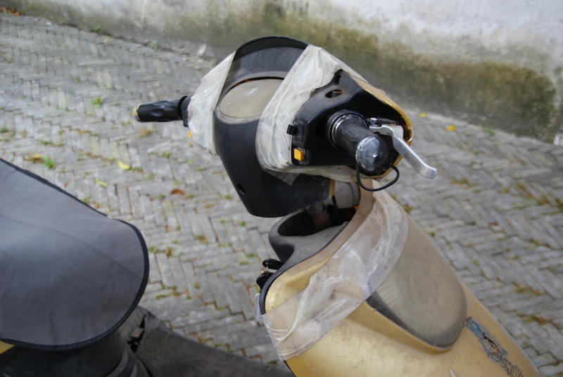 Close view of a scooter handlebar and headlight wrapped in white duct tape.
