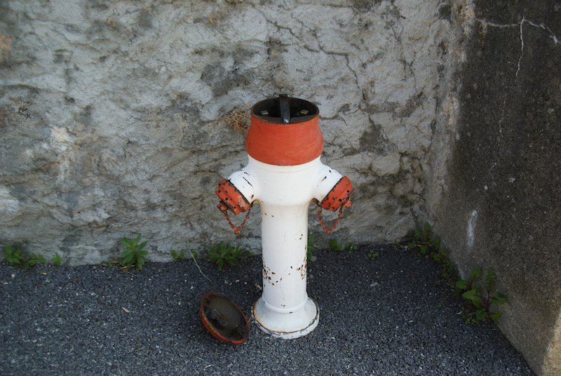 White and red fire hydrant with its top missing and a detached circular cover on gravel beside a stone wall.