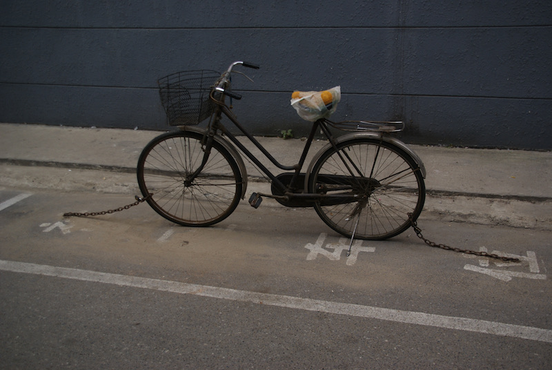 A black bicycle secured with chains in a marked parking space against a dark wall.