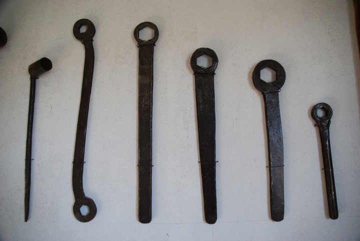 Six dark metal hand tools of different shapes and sizes mounted vertically on a pale wall.