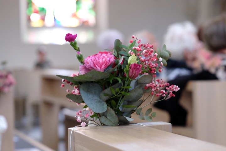 Pink flowers and eucalyptus on a pale wooden church pew, with seated people softly blurred in the background.