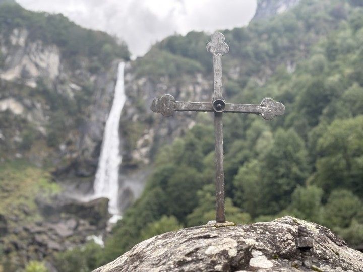 A weathered metal cross fixed to a rock in the foreground, with a tall waterfall descending a green mountainside behind it in Foroglio.