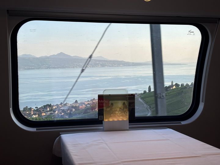 A lake, town and mountain ridge seen through a train dining-car window, crossed by railway wires, with vineyards, a menu stand and a white tablecloth in the foreground.