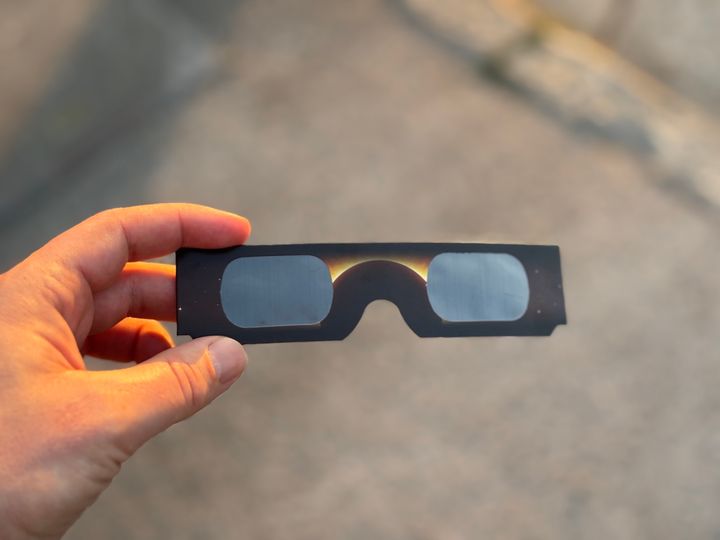 A hand holds black cardboard eclipse glasses over pavement in the warm, dim light of a solar eclipse.