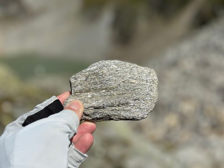 A gloved hand holds a layered grey rock against a blurred rocky landscape.