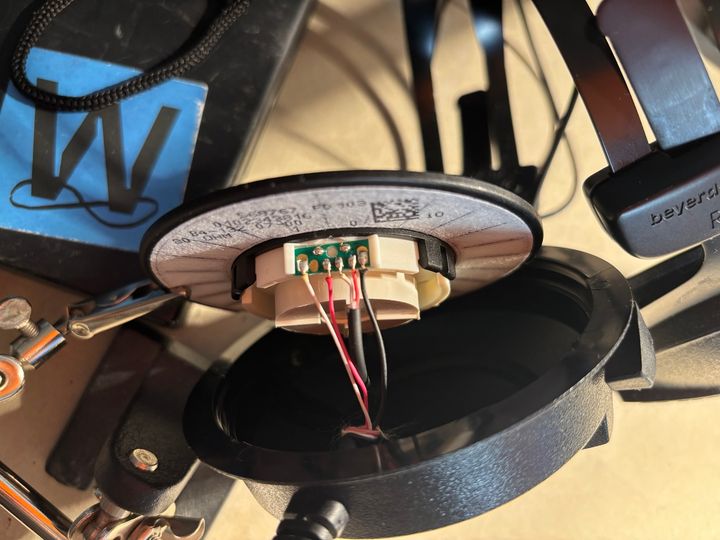An opened headphone earcup with exposed driver, wiring and circuit board beside a helping-hand clamp.